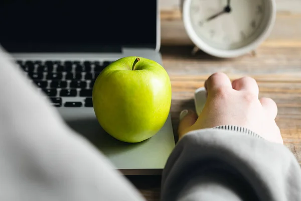 A woman works at a laptop, an apple close-up, an alarm clock on the background, the concept of a healthy breakfast and snack at work.