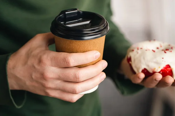 A paper cup of coffee and an appetizing cherry donut in male hands close-up, a white donut in glaze with red sprinkles, a delicious coffee break at work, breakfast.