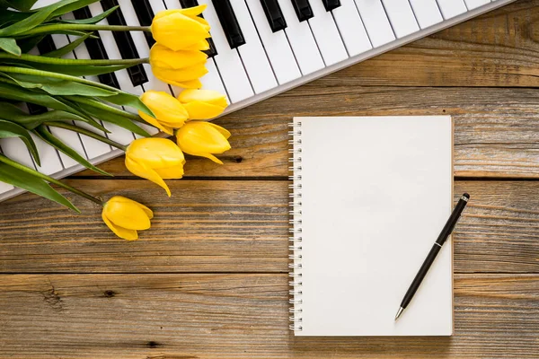 stock image Keys of a white piano, a blank paper notebook and a bouquet of yellow tulip flowers on a wooden background, top view, copy space.