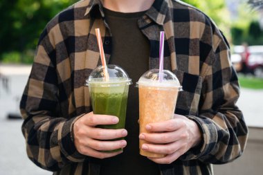 Açık havada elinde yeşil ve turuncu meyve sularıyla plastik bardaklar tutan bir adam. Lezzetli bir yaz içeceği..