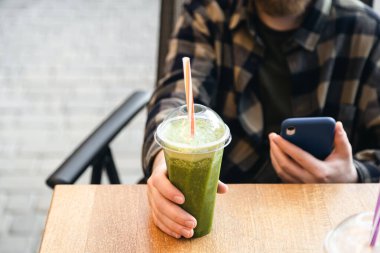 Cafe 'deki masada yeşil meyve püresi. Sağlıklı bir adam diyet protein içeceği içiyor. Elinde plastik bir bardak sebze suyu, sağlıklı yiyecekler, yaşam tarzı..