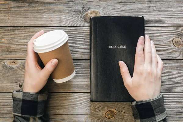 stock image Bible and a paper cup of coffee in male hands on a wooden background, top view, concept of christianity and religion.