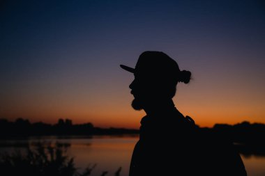 Gün batımında göl kenarında çörekli ve şapkalı sakallı bir adam, bir adamın siyah silueti..