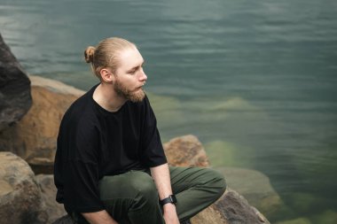 Nehir kenarındaki kayalıklarda sakallı ve çörekli genç bir adam oturuyor..