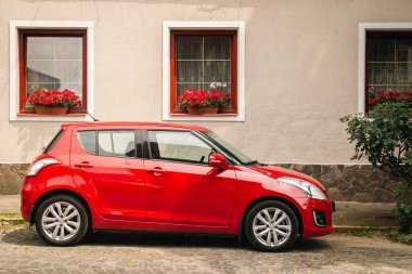 Kırmızı pencereli ve çiçekli eski bir evin önündeki yamaçta modern kırmızı araba, eski Avrupa caddesinde araba..