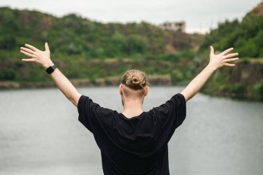 Genç bir adam ellerini kaldırdı, dağlarda bir gölün önünde duruyordu, vahşi doğa, turizm konsepti, arkadan görüş..