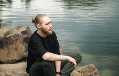 Nehir kenarındaki kayalıklarda sakallı ve çörekli genç bir adam oturuyor..