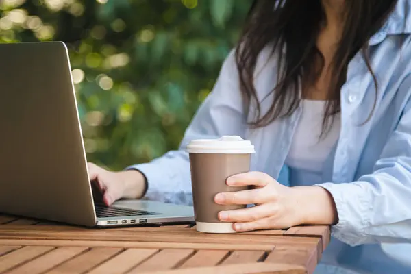 A woman with a paper cup of coffee works at a table with a laptop outside on a blurred background of nature.