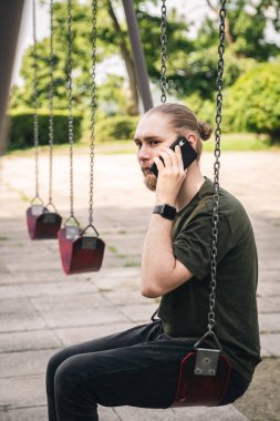 Sakallı genç bir adam parkta salıncakta sallanıyor ve telefonda konuşuyor..