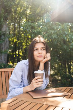 Bir fincan kahve ve dizüstü bilgisayarı olan genç bir kadın dışarıda terasta tahta bir masada otururken güneşin tadını çıkarıyor..