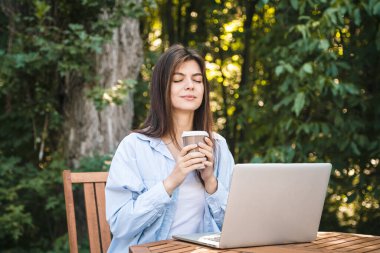 Dizüstü bilgisayarı ve bir fincan kahvesi olan genç bir kadın ormanın arka planında bir masada oturur ve açık hava, ruh sağlığı ve tatil kavramının tadını çıkarır..