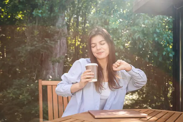 Happy satisfied laptop user woman relaxing with cup of coffee, stretching body with sleepy closed eyes, rising hands, breathing fresh air outside.