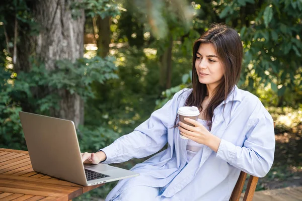 Attractive young woman with a cup of coffee works outside with a laptop, sits at a wooden table on the background of the forest, the concept of working on vacation, freelancing and studying online.