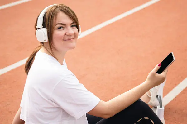 Kulaklıklı bir kadın stadyumdaki koşu bandında akıllı telefon kullanıyor, antrenmandan sonra dinleniyor veya koşuyor, fitness uygulamaları kullanıyor, dijital kavram kullanıyor..