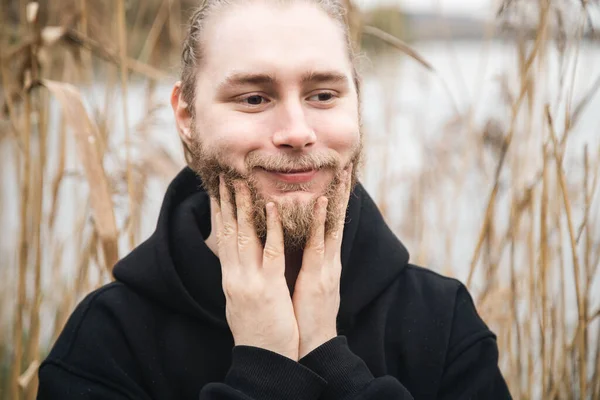 Grooming, skin care and man concept, smiling young bearded caucasian man touching his face on reeds background.