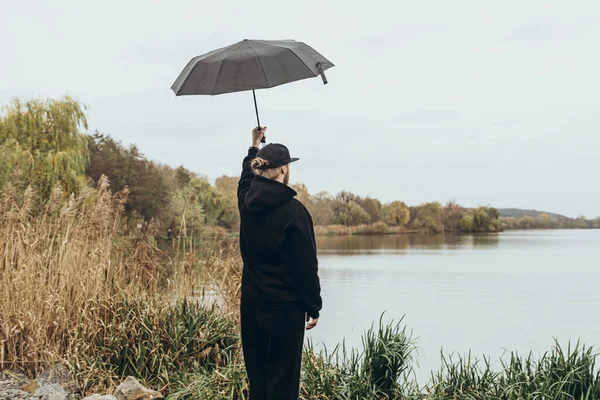 A stylish young man in gloomy weather by the river raises his hand with an umbrella high up, view from the back.
