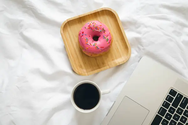 Pink donut, cup of coffee and laptop in white bed, top view, home office, breakfast in bed.