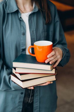 Bir kadın kütüphanenin arka planında bir yığın kitap ve turuncu bardak tutuyor. Bir yığın kitap ve bir kupa kadının elinde. Eğitim, öğrenim ve okuma konsepti.