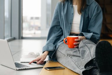 Turuncu bardaklı genç bir kadın dizüstü bilgisayar kullanıyor. Bir öğrenci ödevini yapıyor ve çevrimiçi çalışıyor. Uzak çalışma ya da serbest çalışma. Boşluğu kopyala.