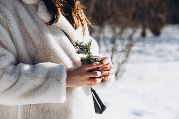 A woman in a white fur coat with a paper glass of a hot drink in the forest. Outdoors winter activities. Winter Christmas and lifestyle concept. Copy space.