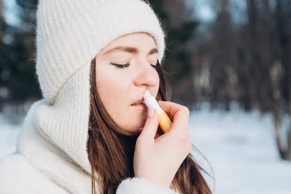Woman applying lip balm outdoors in winter. Portrait of a beautiful woman with application of the protective cream to the lips. Chapped, or cracked, dry lips that have lost moisture.