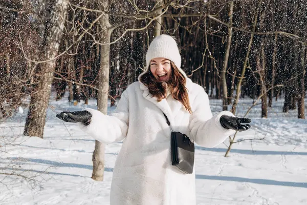 A beautiful woman in winter clothes is enjoying the winter. Happy woman in a white fur coat and hat in a winter forest in sunny weather. Outdoors winter activities.