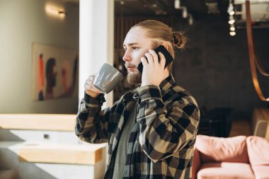 Genç sakallı bir adam modern bir ofiste kahve içer ve telefonda konuşur. İşte kahve molası. Teknoloji ve iletişim iş başında. Boşluğu kopyala.