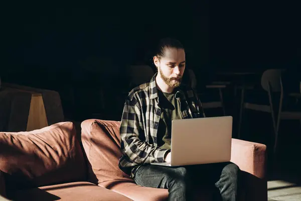 A bearded young man works in a cafe with a laptop remotely. An IT specialist sits on the couch on a call in the morning. Young student in a cafe studying online. Copy space.