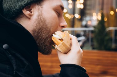 Genç sakallı bir adam iştah açıcı bir şekilde Bokeh 'in bulanık arka planında hamburger yiyor. Aç bir adam hamburgeri ısırır, yakın plan..