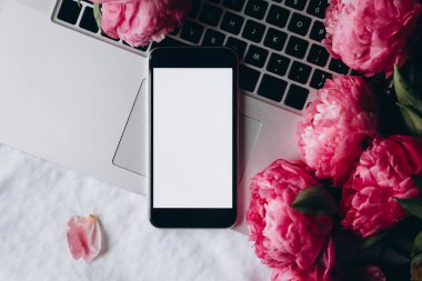 Beyaz ekranlı akıllı telefon, dizüstü bilgisayar ve yatağın üstünde pembe şakayık çiçekleri, üst manzara. Akıllı telefon ve çiçeklerle bahar arkaplanı, boşluğu kopyala.