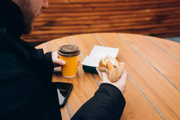 Bir adam hamburger yer ve kahvesini içerken dışarıdaki bir fast food kafenin ahşap masasında oturur. Yaşam tarzı fotoğrafı, sağlıksız yemek konsepti, abur cubur..