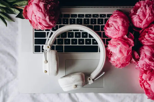 White headphones, laptop and pink peony flowers on white sheets in bed, concept of remote work, music creative. Flat lay, top view spring minimalist home workspace.
