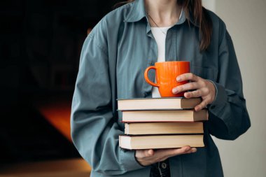 Bir kadın kütüphanenin arka planında bir yığın kitap ve turuncu bardak tutuyor. Bir yığın kitap ve bir kupa kadının elinde. Eğitim, öğrenim ve okuma konsepti.