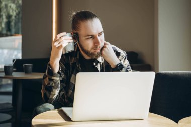Uykulu bir adam bir fincan kahveyle sabahleyin bir kafede dizüstü bilgisayarın önünde oturur. Yorgun öğrenci çevrimiçi çalışıyor. İş yerinde yanma.