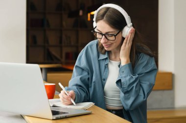 Uzaktan eğitim. Gülümseyen bir kadının portresi, dizüstü bilgisayar kullanarak ve deftere yazarak, not alarak, ders, konferans ya da webinar izleyerek, çevrimiçi çalışarak. Boşluğu kopyala.