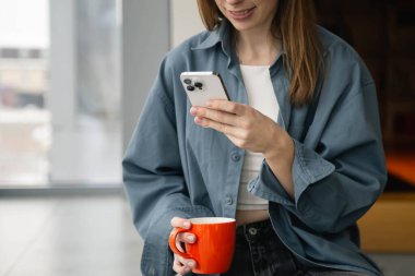 Mavi gömlekli genç bir kadın akıllı telefon kullanıyor ve pencerenin yanında otururken turuncu bir bardak tutuyor. Kahve molası konsepti, işte rahatlama. Boşluğu kopyala.
