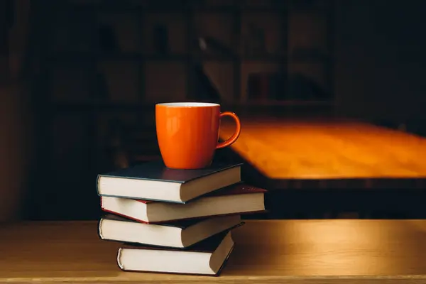 An orange cup on a stack of books on a dark library background. Book background. Education, learning and reading concept. Copy space.