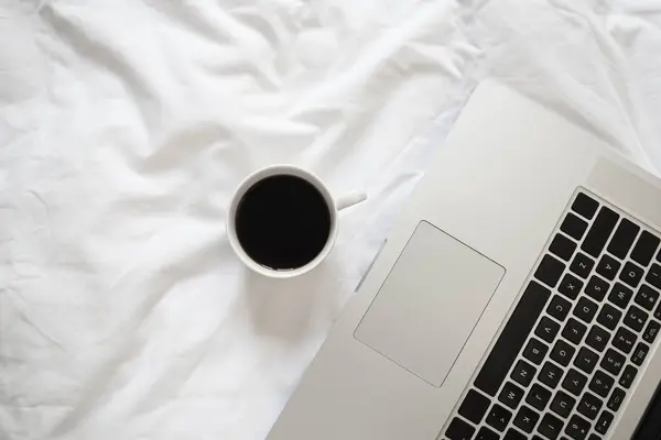 Laptop and coffee in bed on white linens, flat lay, top view minimalist workspace background, working at morning concept.