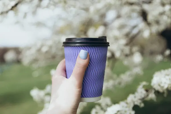 Disposable blue paper cup of coffee in the hand of a girl on a background of white blooming spring tree. Springtime. Beautiful nature scene with blooming tree.