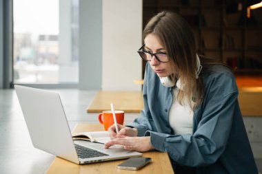 Bir öğrenci not defterine yazar ve laptop ekranına bakar. Gözlüklü genç bir kadın dizüstü bilgisayarın önünde oturur ve ders çalışır. Eğitim kavramı, çevrimiçi ve uzaktan öğrenme.