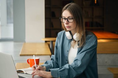 Bir öğrenci not defterine yazar ve laptop ekranına bakar. Gözlüklü genç bir kadın dizüstü bilgisayarın önünde oturur ve ders çalışır. Eğitim kavramı, çevrimiçi ve uzaktan öğrenme.