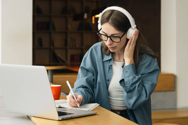 Distance education. Portrait of smiling woman sitting at desk, using laptop and writing in notebook, taking notes, watching tutorial, lecture or webinar, studying online. Copy space.