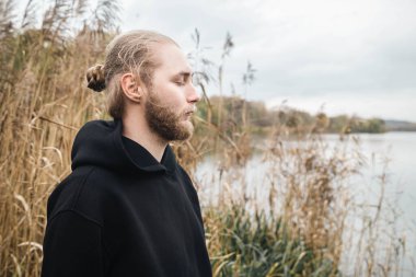 Genç, sakallı, sarışın bir adam gözleri kapalı olarak kıyıda duruyor. Ön tarafta sazlıklar, akıl sağlığı, açık hava eğlencesi, kopyalama alanı..