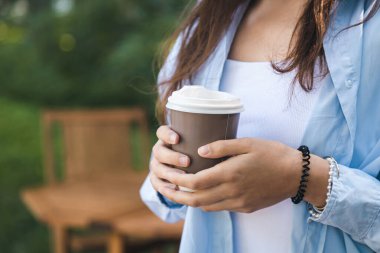 Mavi tişörtlü genç bir kadın elinde tek kullanımlık bir bardakla doğanın arka planında kahve tutuyor..
