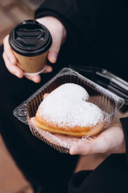 Bir fincan kağıt kahve ve erkek ellerinde kalp şeklinde bir çörek, yakın plan. Dışarıda donutu olan siyah ceketli bir adam..