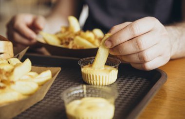 Yakından bakınca, bir adam patates kızartması yiyor. Yemek konseptini aldatmak. Yemek, yaşam tarzı konsepti. Bir erkek eli patates kızartmasını tutar ve peynir sosuna batırır. Çıtır patatesler derinde.