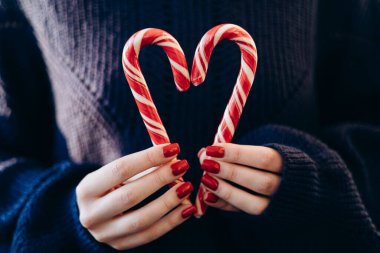 Elinde Noel şeker kamışı tutan kadın, yaklaşın. Kalp şeklinde iki şeker kamışı. Hayırseverlik kavramı, iki şeker tutan kadının göğüs seviyesinde kalp oluşturması. Çocuklar için tatlı karamel. Rahat süveter.