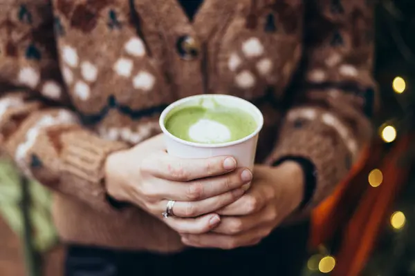 Kışın bir kafe yakınında elinde matcha latte tutan bir kadın. Elinde, bulanık Noel ağacı arkaplanlı bir kibrit içeceği tutan kadın. Noel kış tatili konsepti.