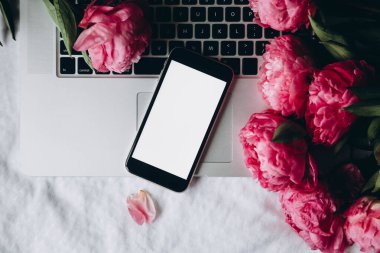 Beyaz ekranlı akıllı telefon, dizüstü bilgisayar ve yatağın üstünde pembe şakayık çiçekleri, üst manzara. Akıllı telefon ve çiçeklerle bahar arkaplanı, boşluğu kopyala.