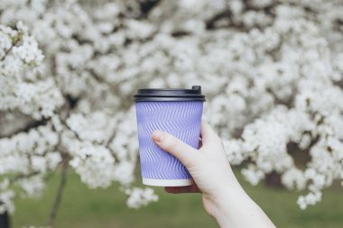 Beyaz çiçek açan bahar ağacının arka planında duran bir kızın elinde tek kullanımlık mavi kahve fincanı. İlkbahar. Çiçek açan ağaçlı güzel doğa sahnesi.